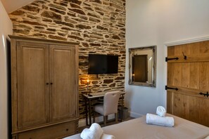 A room with a stone wall, wooden wardrobe, desk with chair, bed with towels, TV, and a mirror. - The Cart Shed (St Issey)