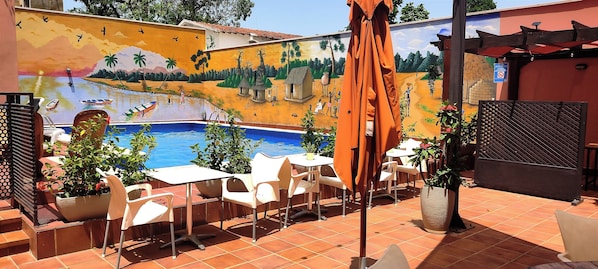 Un patio exterior con piscina, sillas blancas y un mural en la pared.