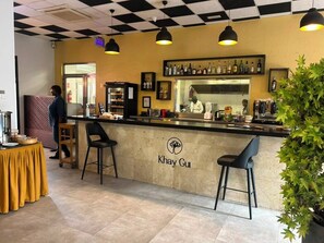 Interior de restaurante con techo a cuadros, barra con taburetes, zona de cocina y área de comedor con mantel amarillo.