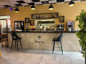 Zona de bar con mostrador, dos taburetes, un chef al fondo y una pared con un logotipo.