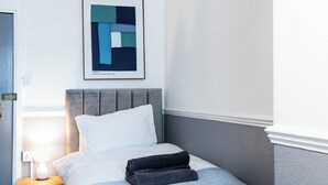 En pænt redt seng med et gråt sengegavl, et natbord med en lampe, og et indrammet abstrakt kunstværk på væggen.