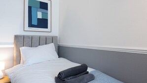 En pænt redt seng med en pude, et sammenrullet håndklæde og et indrammet abstrakt billede på væggen.