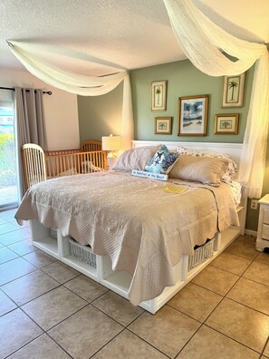 A bedroom with a bed, curtains, a crib, a nightstand, and wall art. - Young Avenue Retreat -Steps to Cocoa Beach (Cocoa Beach)