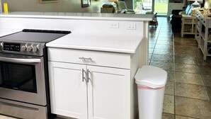 Une cuisine équipée d’une cuisinière, d’un four et de meubles blancs. On y trouve également un ventilateur de plafond, une poubelle et une fenêtre donnant sur l’extérieur.
