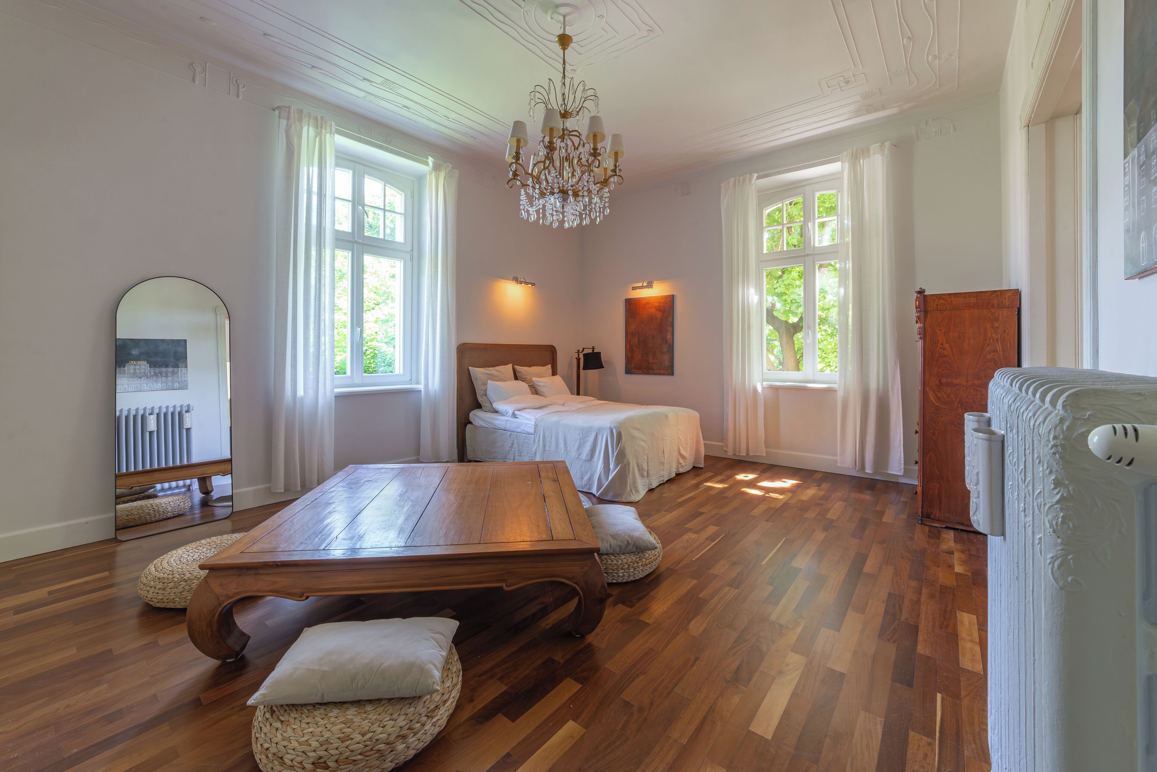 Une pièce avec un plancher en bois, une grande table en bois, deux lits, un lustre et de grandes fenêtres.