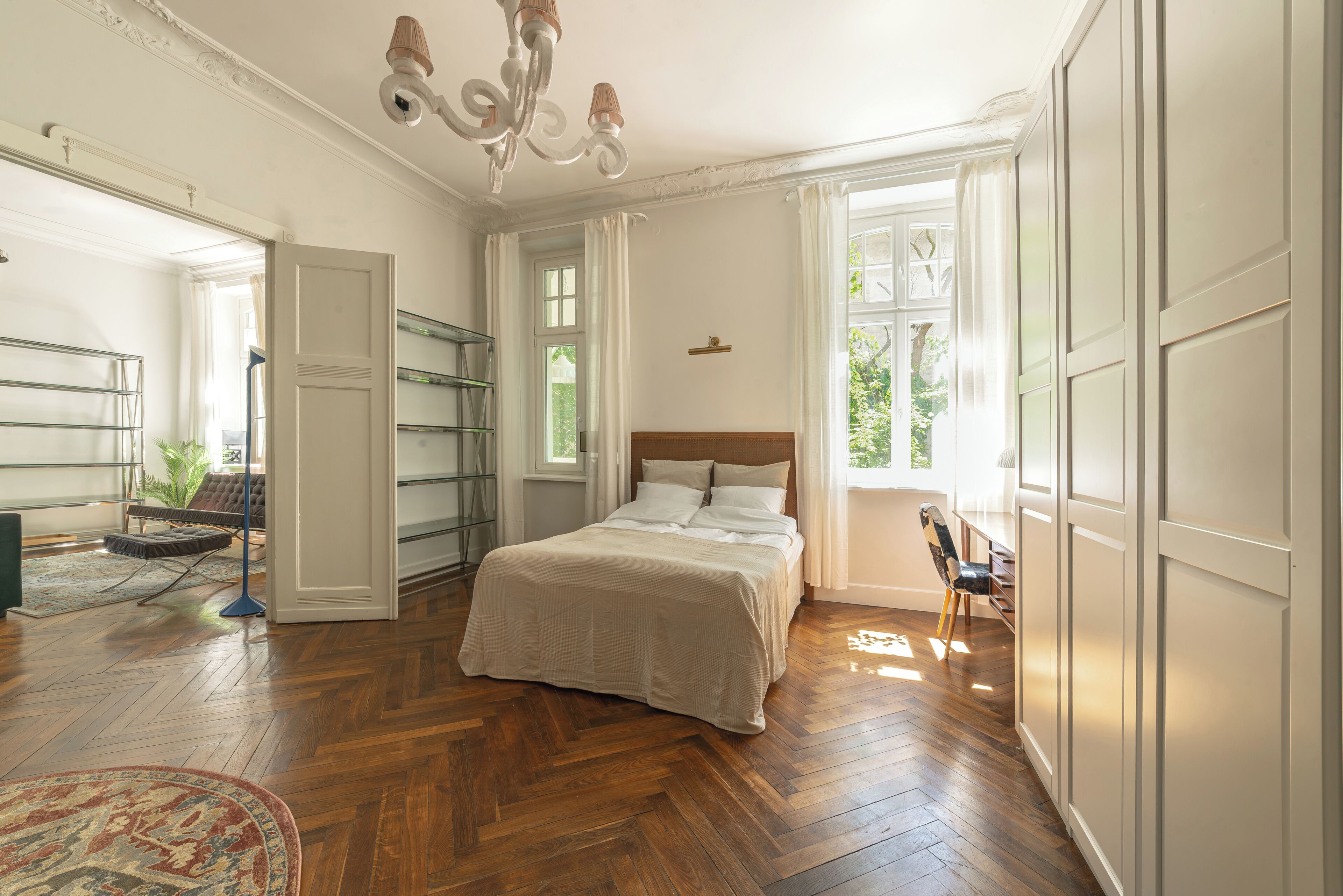 Une chambre avec un plancher en bois, un lit recouvert de draps blancs, un lustre et une vue sur la verdure à l’extérieur.