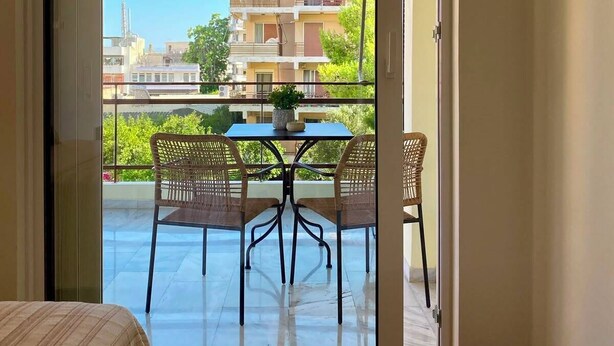 A room with a bed, a table with chairs, and a sliding glass door leading to a balcony with a view of buildings and greenery. - Cosy apartment In the heart of Varkiza 100 meters from the Sea and all Shop area (Vari)