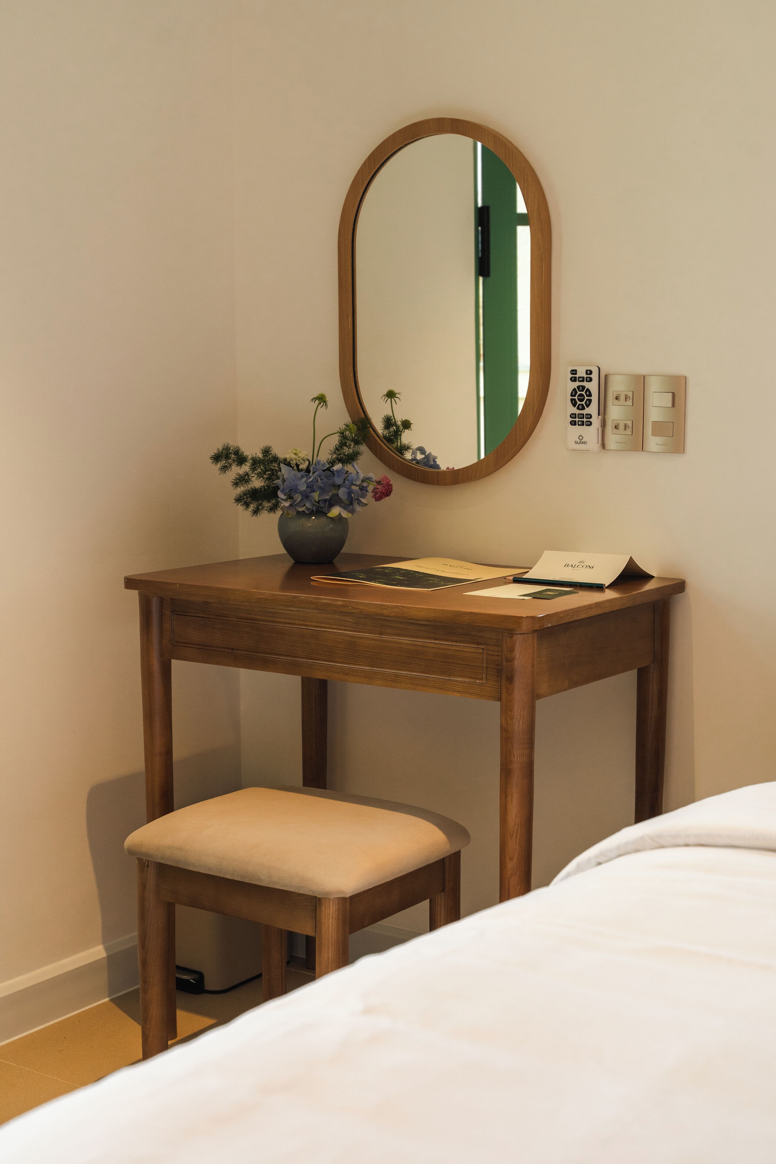 A wooden bedside table with a mirror, a small stool, and a vase with flowers.