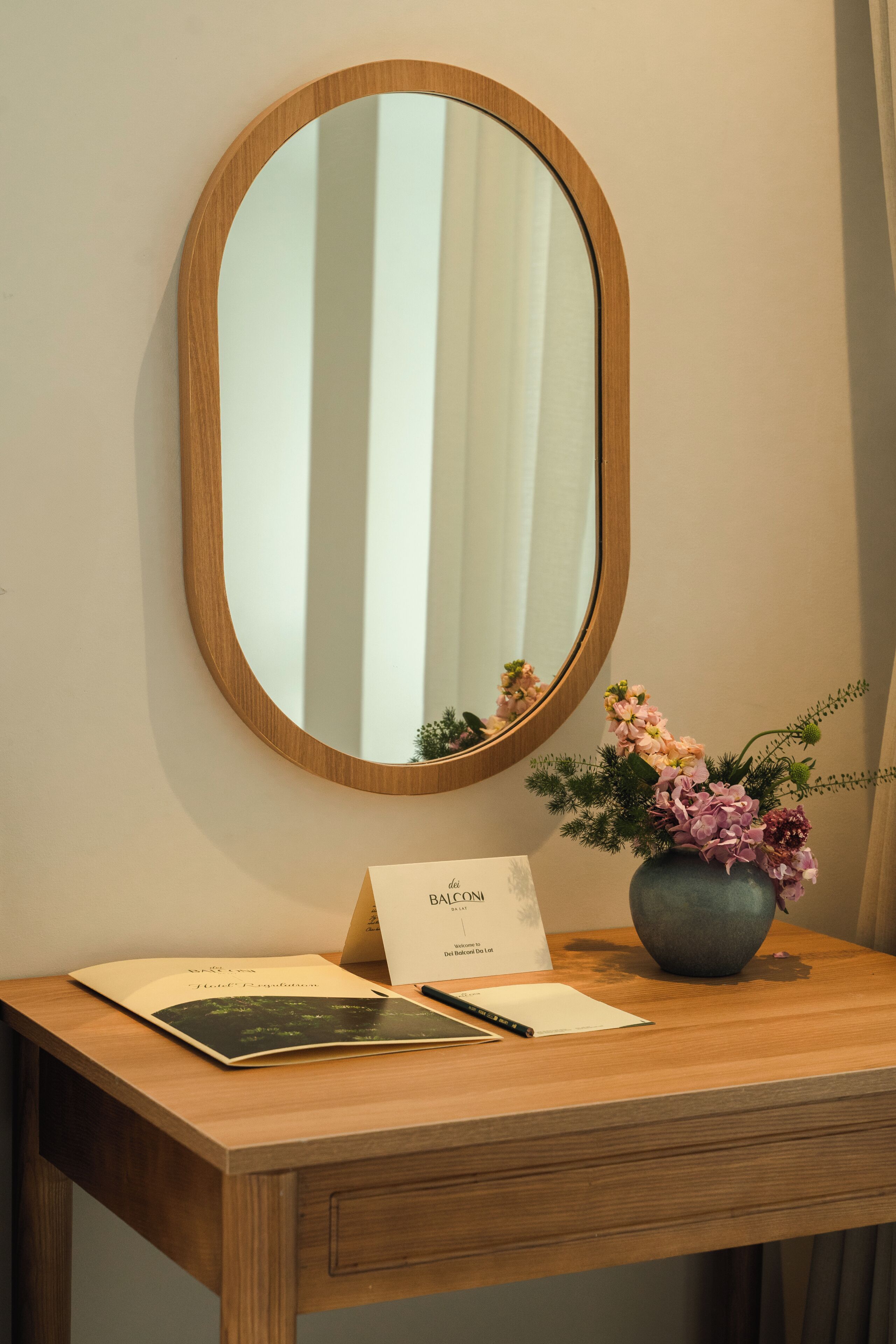 A wooden desk with a mirror, a vase with flowers, and a welcome card.