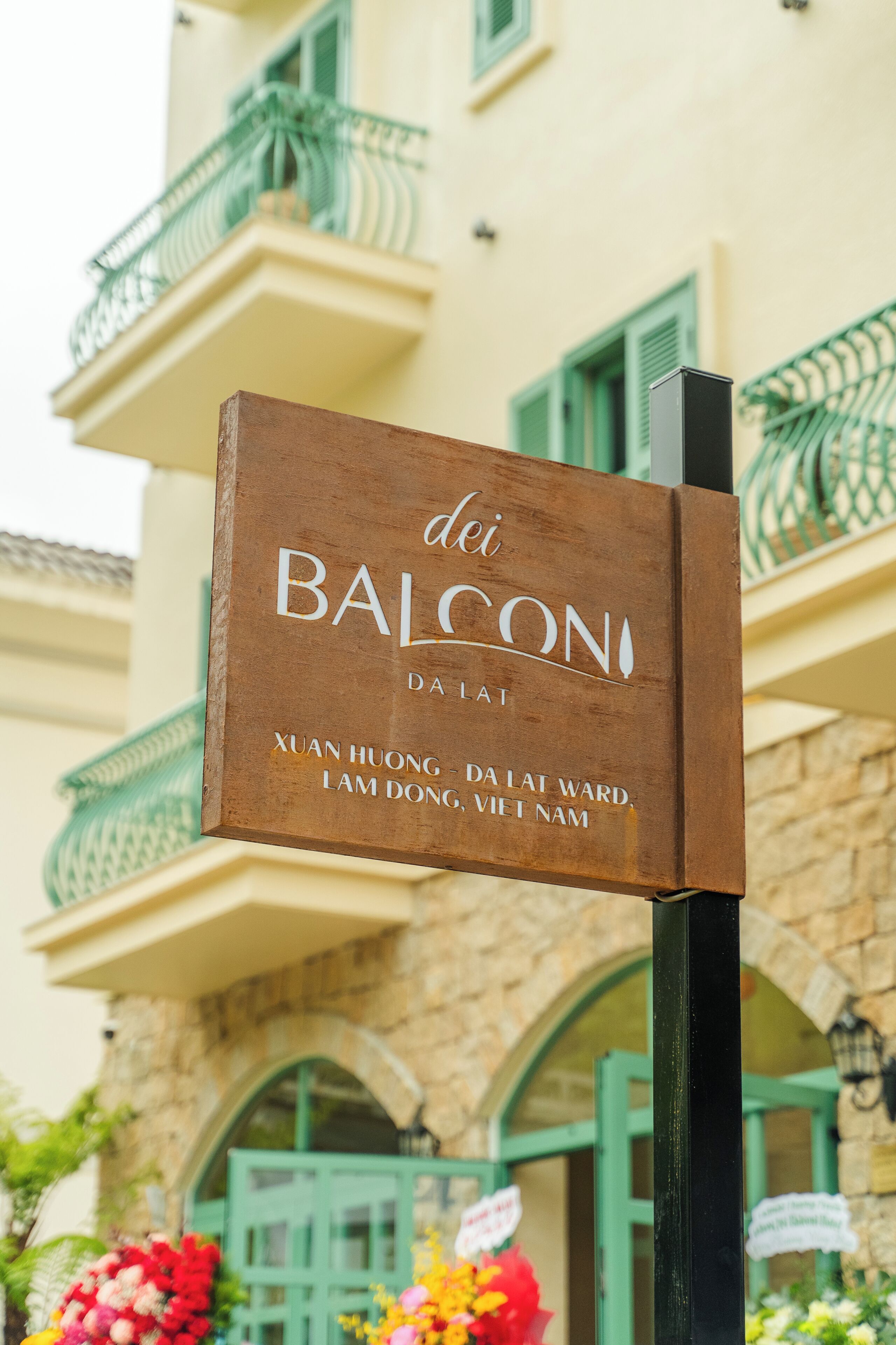 Un letrero que dice 'Balcon da Lat', con un edificio de piedra y balcones verdes al fondo.