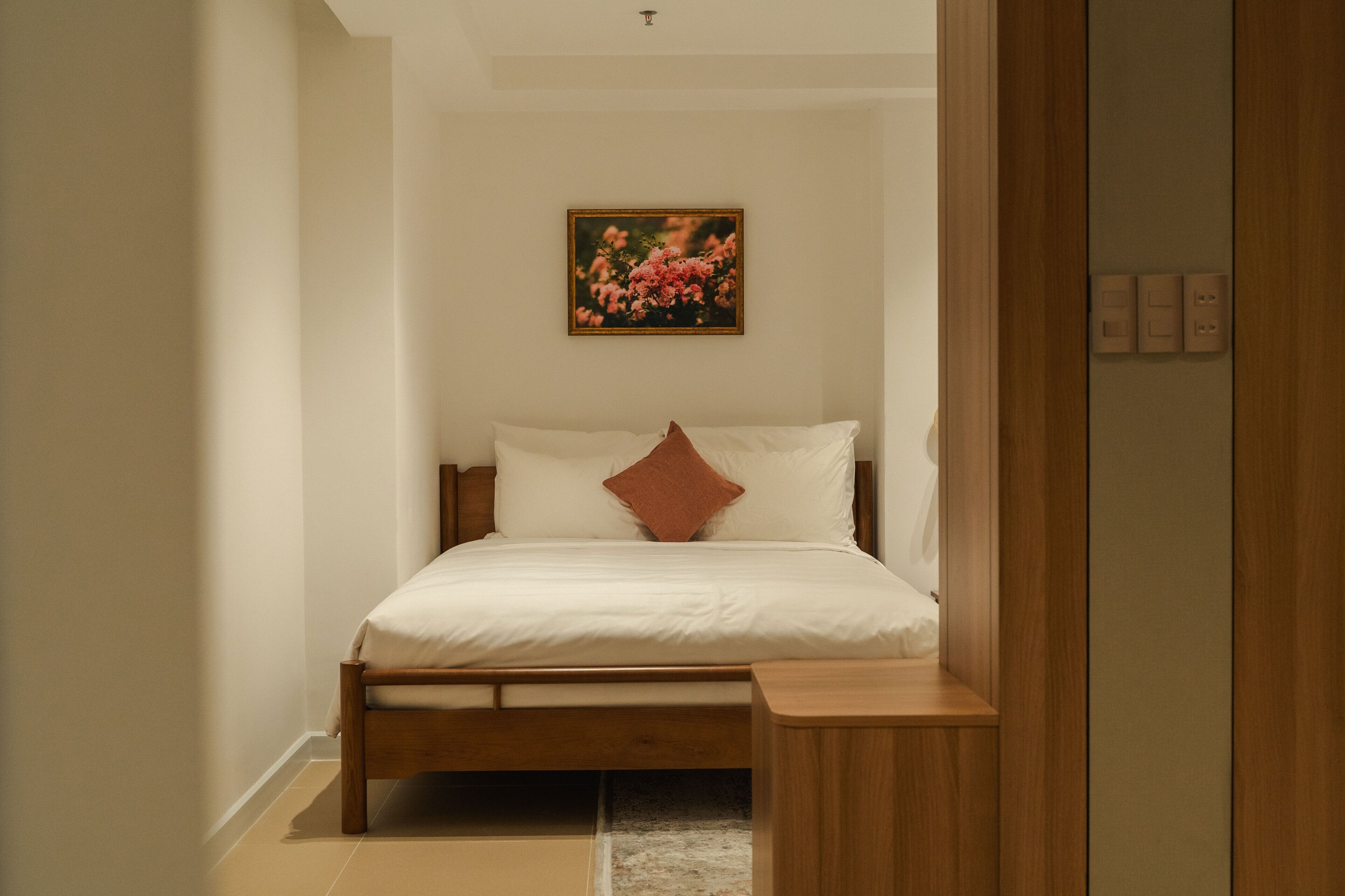 A bedroom with a wooden bed, white bedding, a single brown pillow, a small wooden nightstand, and a framed picture on the wall.