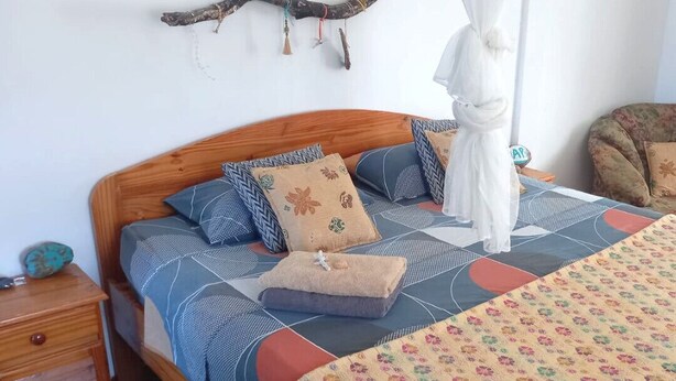 Un dormitorio con cama de madera, varios almohadones, una rama decorativa en la pared y una mesita de noche con lámpara.