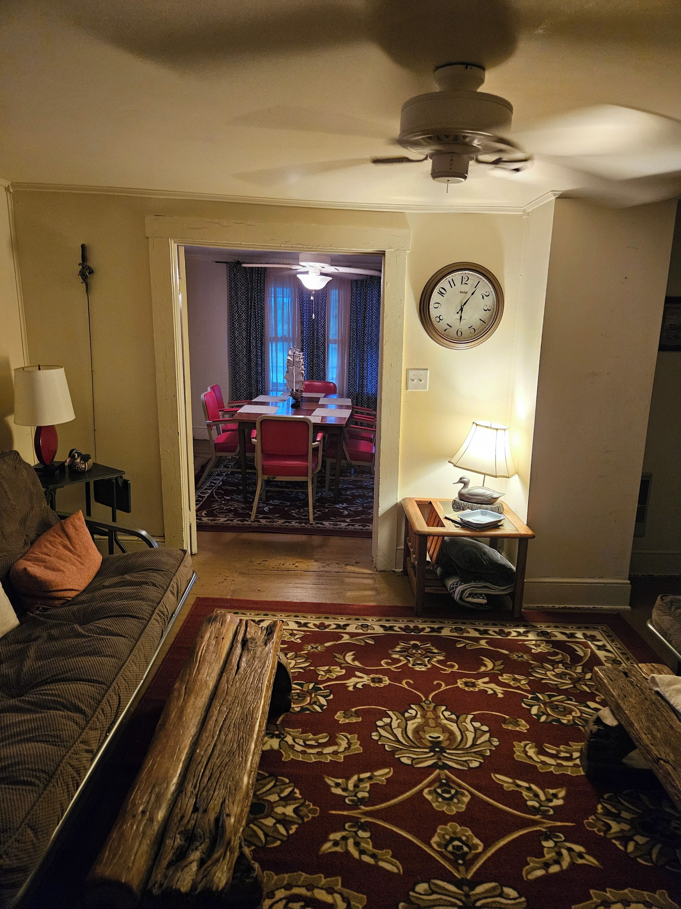 A living room with a sofa, a coffee table, a rug, a dining area with a table and chairs, and a clock on the wall.