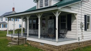 A white house with a green roof, a porch with chairs, and a visible chimney.