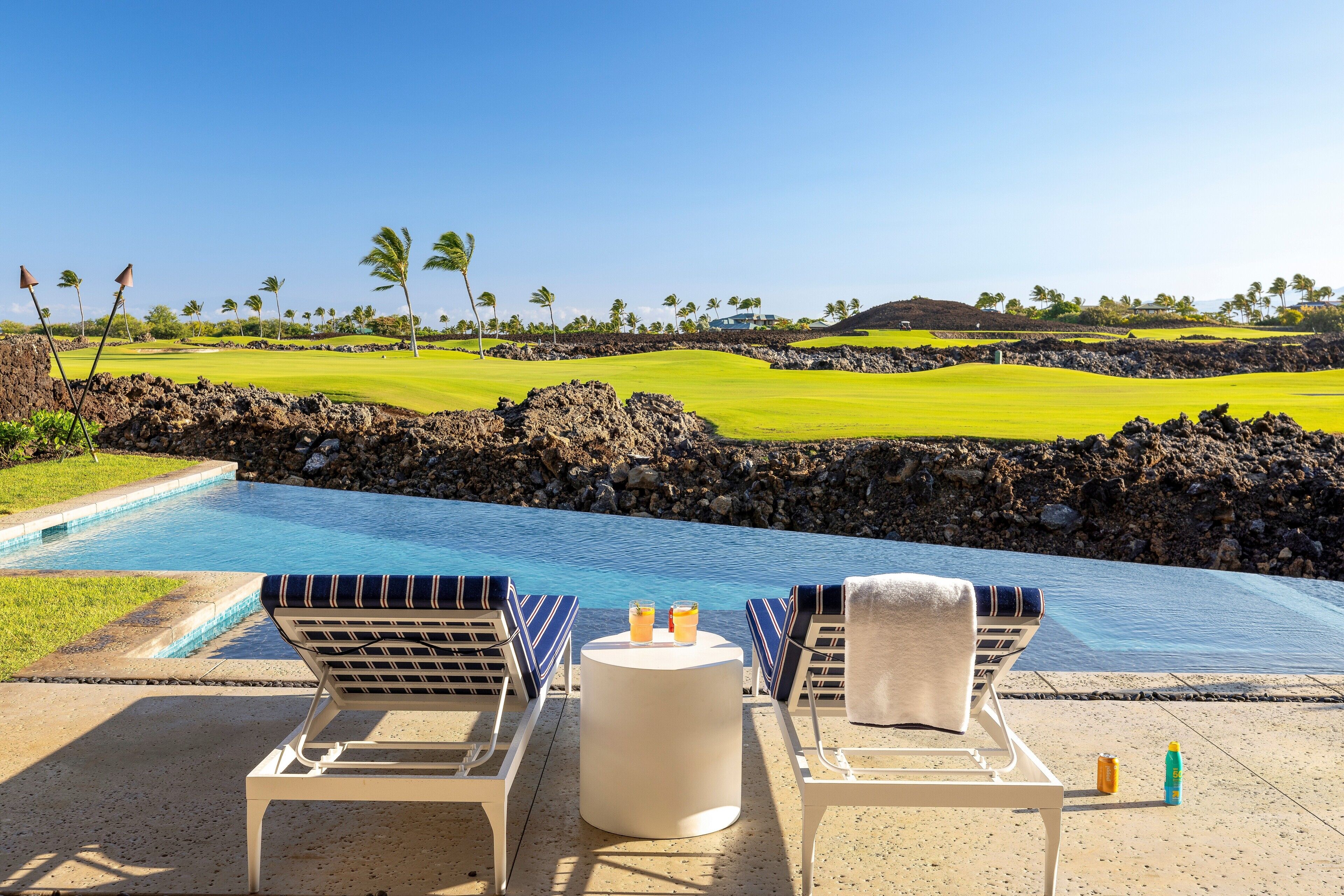A poolside area with lounge chairs, a table with drinks, and a view of a golf course and ocean.