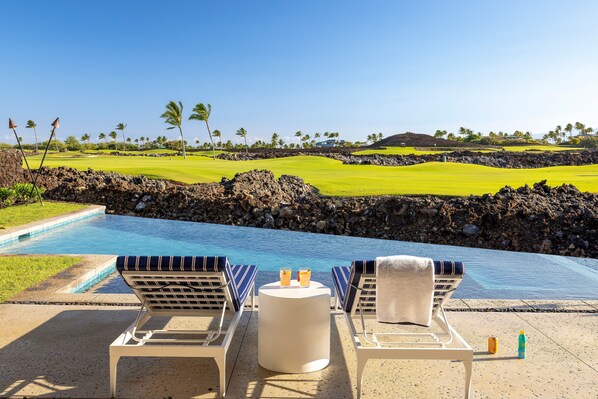 Área da piscina com espreguiçadeiras, uma mesa com bebidas e vista para um campo de golfe e o oceano.