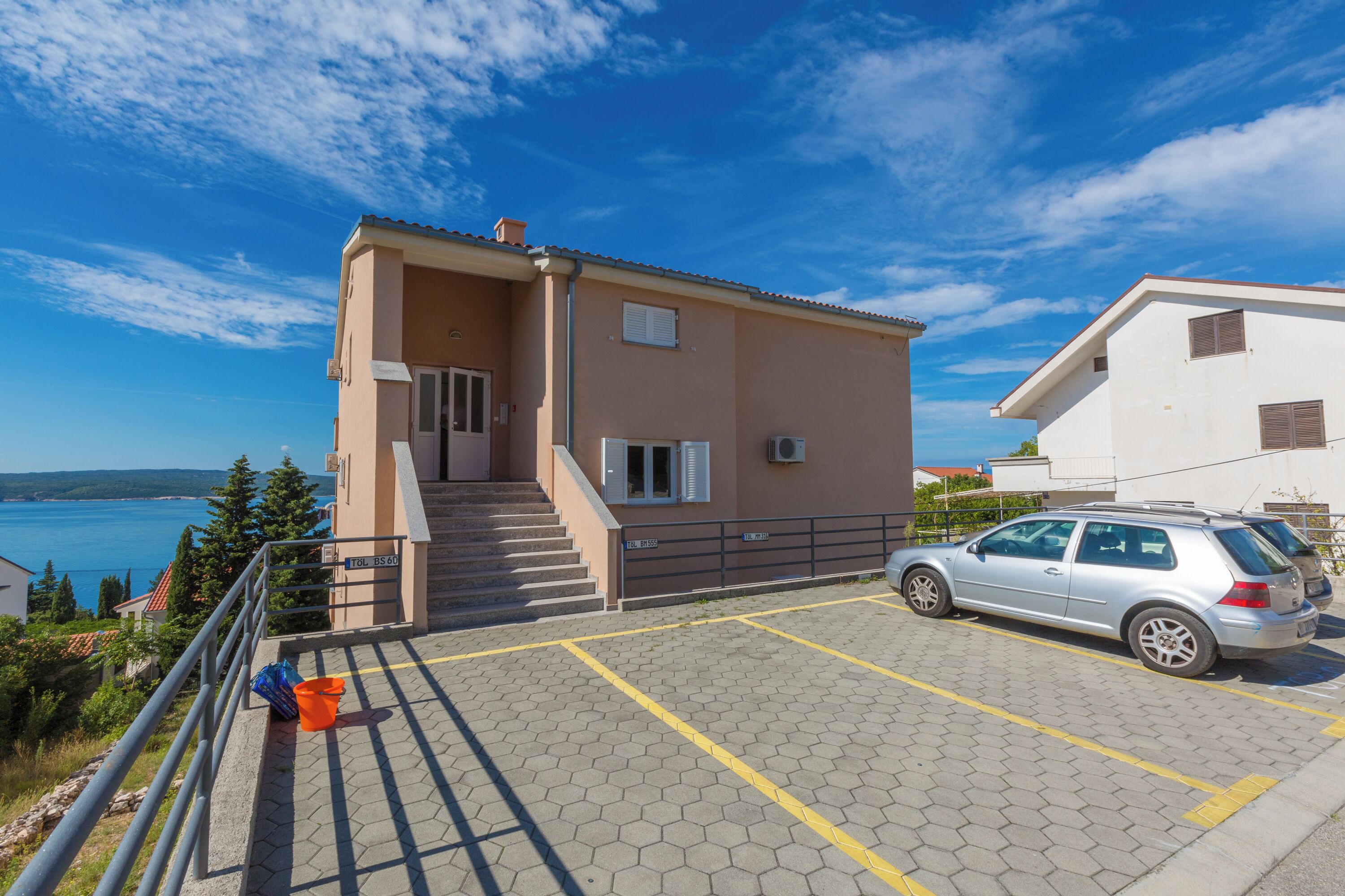 Ein Gebäude mit einer Treppe zu einer Tür, ein Parkplatz mit einem Auto und Blick aufs Meer.