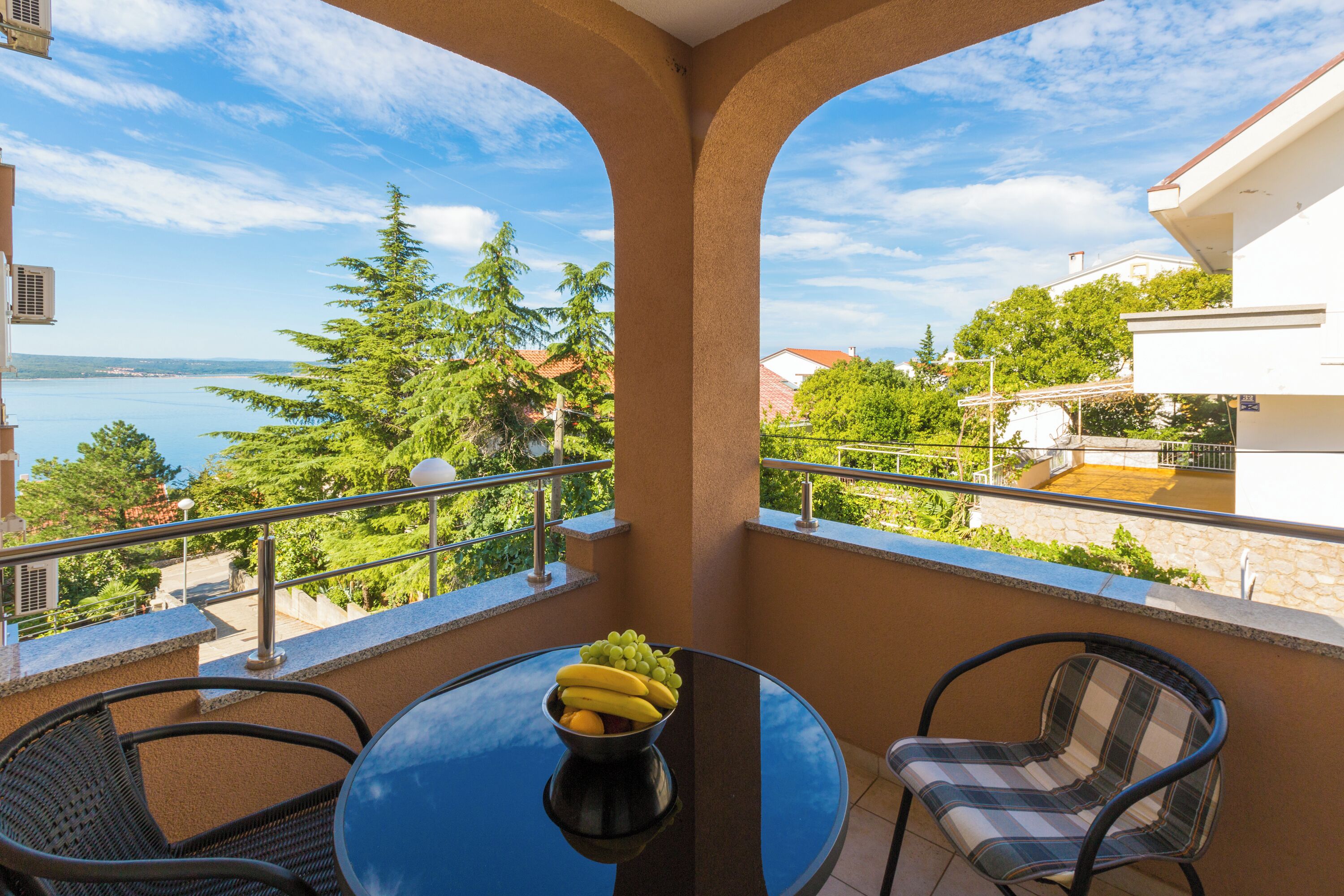 Un balcone con tavolo e sedie, una ciotola di frutta e vista sul mare e sugli alberi.