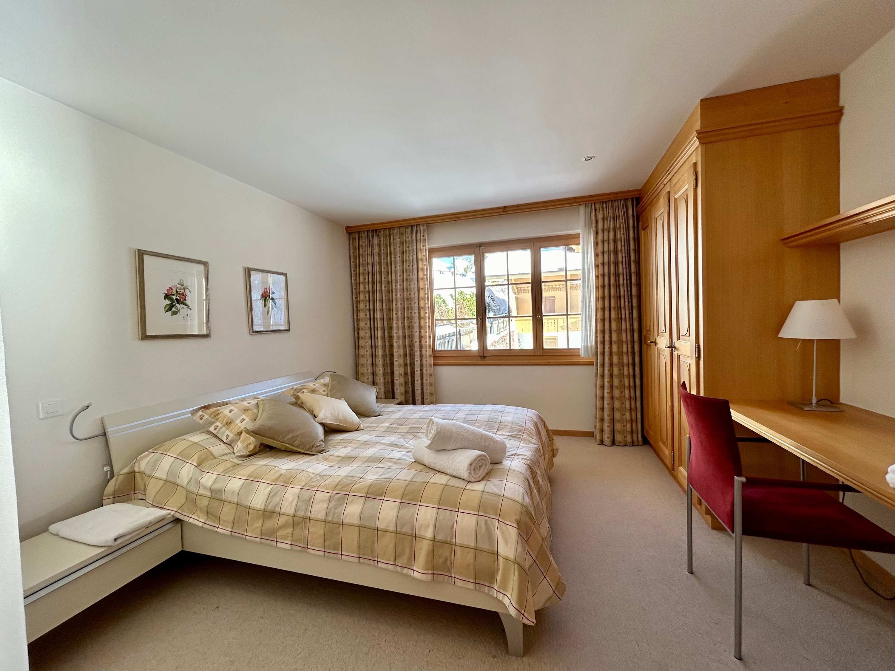 A bedroom with a bed, a desk, a chair, a window with curtains, and two framed pictures on the wall.