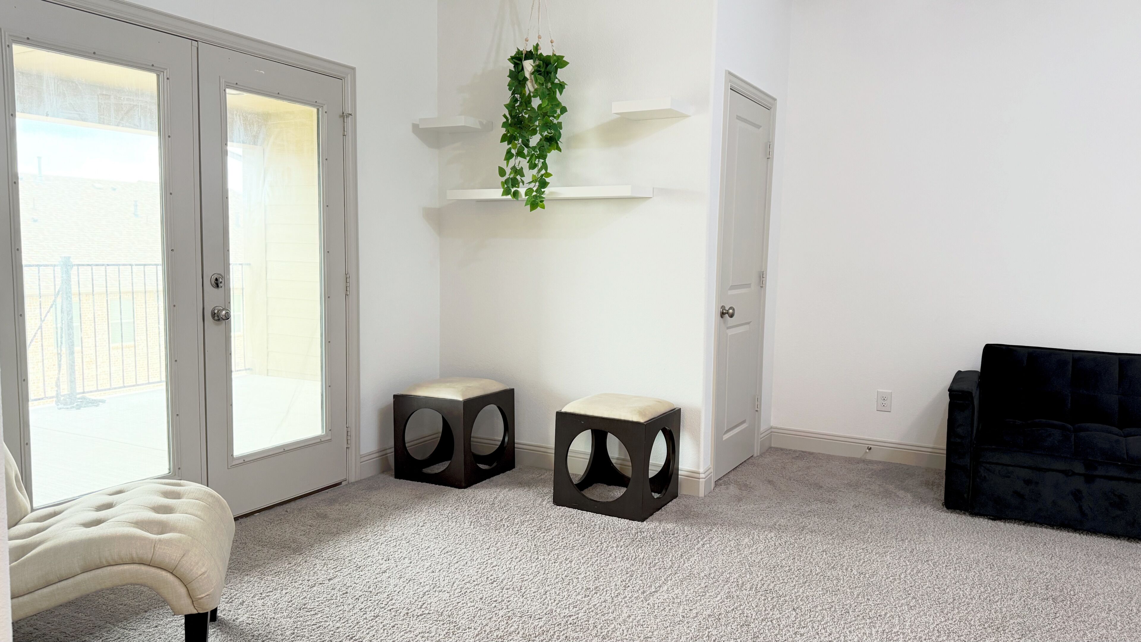A room with a bed, two stools, a hanging plant, and a black sofa.
