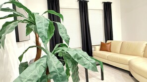 A living room with a beige sofa, a large green plant, black curtains, and a window with blinds. - Stylish Frisco Retreat Near The Star & Hotspots (Frisco)