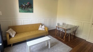 Ein Wohnzimmer mit gelber Couch, einem weißen Couchtisch, einem Esstisch mit Stühlen, einem Bild an der Wand und einer dekorativen Deckenleuchte.
