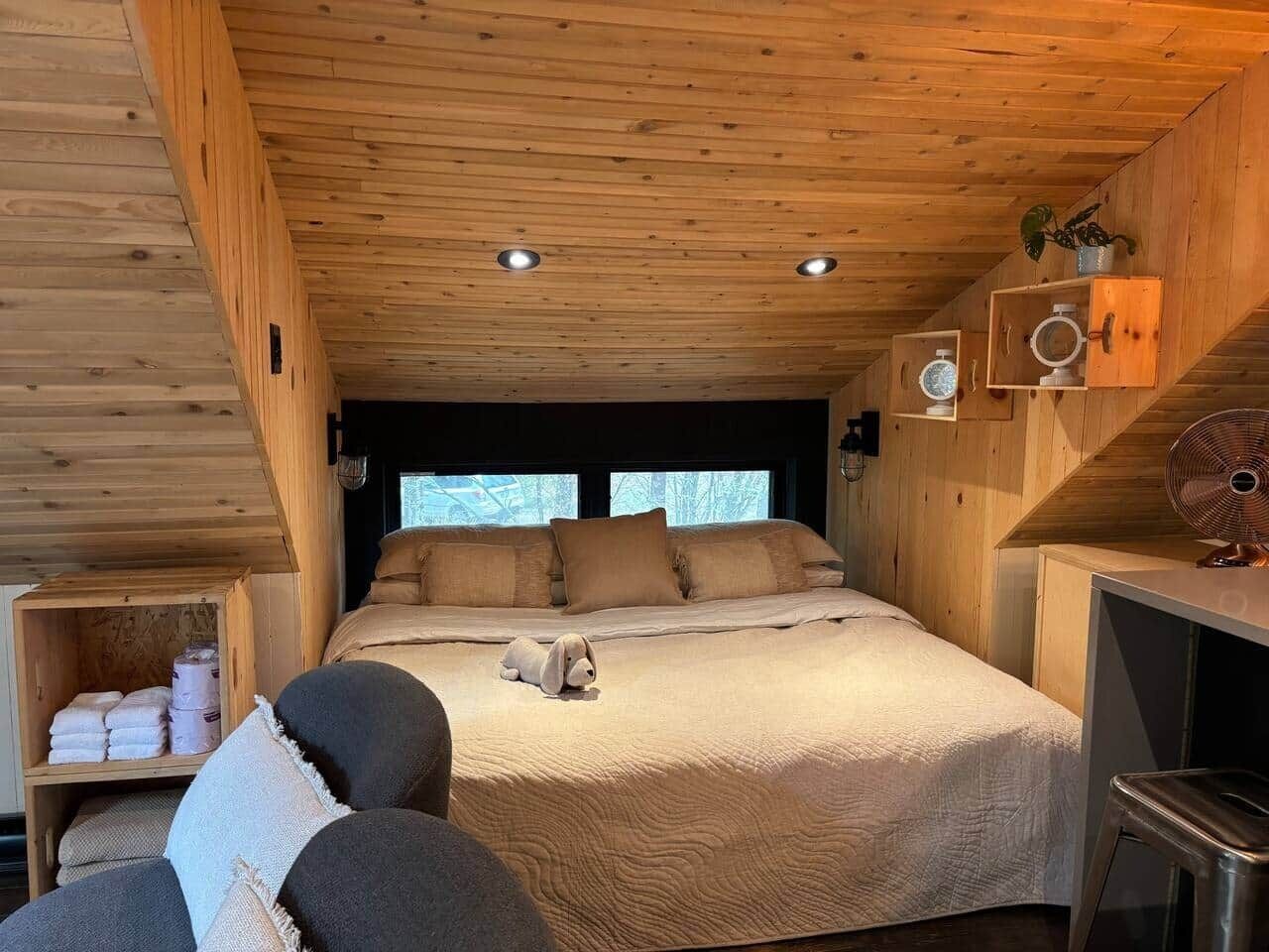 A wooden cabin bedroom with a bed, pillows, a small table, a chair, and a shelf with towels.