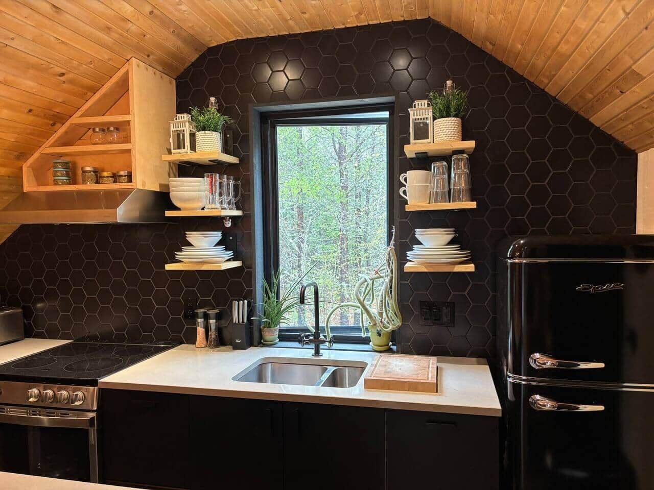 Une cuisine avec un dosseret hexagonal noir, des étagères en bois, un réfrigérateur noir et une fenêtre donnant sur des arbres.