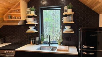 Une cuisine avec un dosseret hexagonal noir, des étagères en bois, un réfrigérateur noir et une fenêtre donnant sur des arbres.