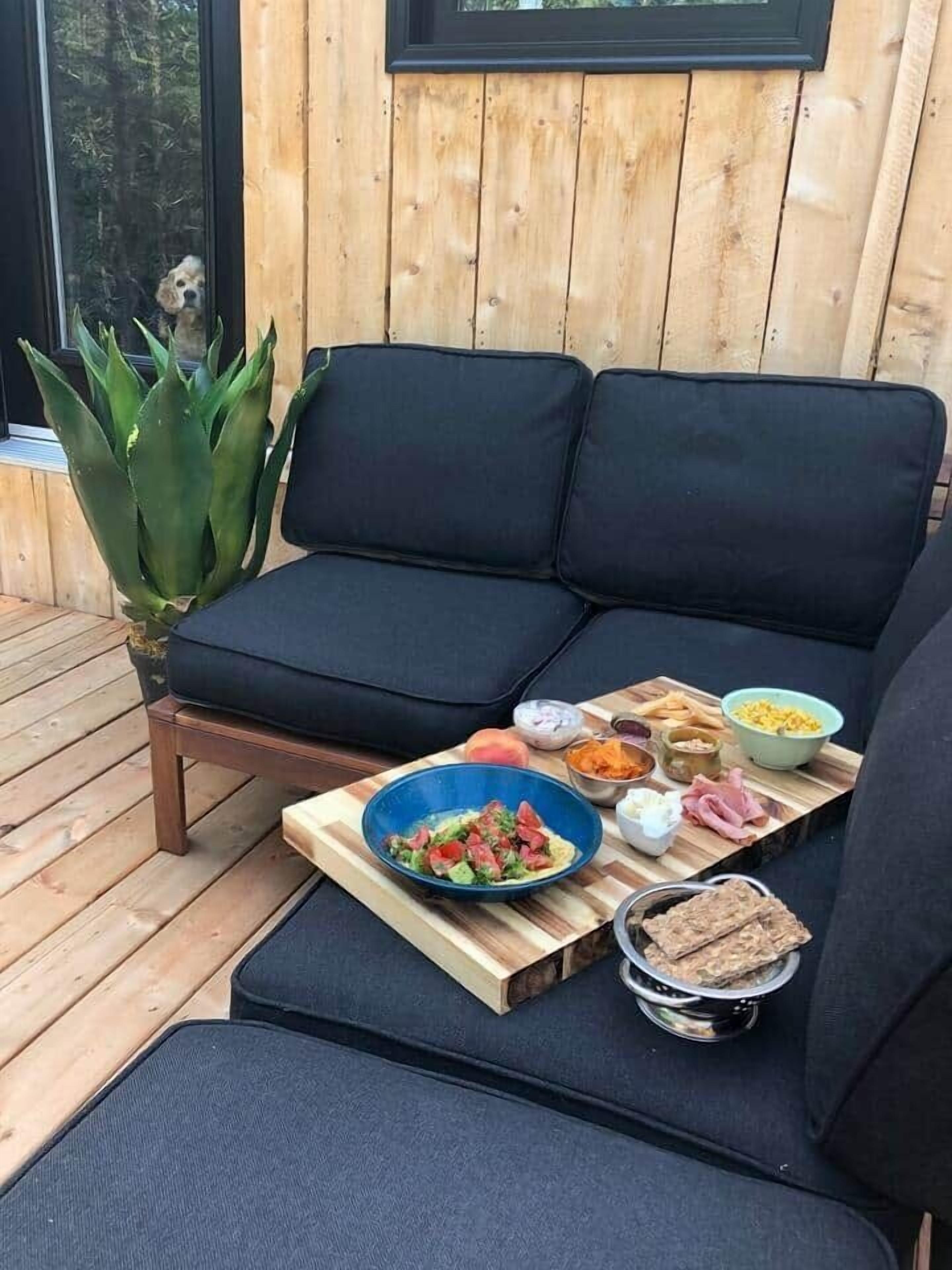 Una terraza de madera con un sofá acolchado negro, una mesa de comedor con diferentes alimentos y una planta en maceta.