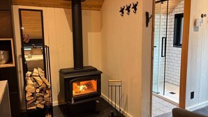A room with a wood-burning stove, a glass door leading to a shower area, and a stack of firewood. - Chalets du plateau des Hautes-Gorges : Le Grenier (Saint-Aimé-des-Lacs)