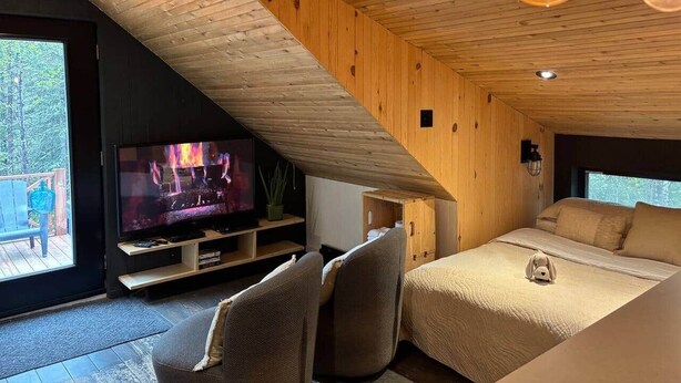 A cozy room with a TV, a bed, and a window overlooking a deck. - Chalets du plateau des Hautes-Gorges : Le Grenier (Saint-Aimé-des-Lacs)
