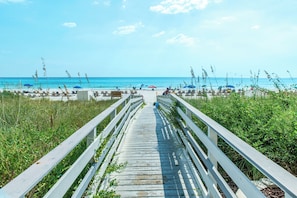 Ein hölzerner Steg führt zu einem Strand mit Sonnenschirmen und Liegestühlen.