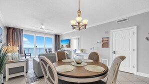 A dining area with a round table, chairs, a chandelier, and a view of the sea through large windows. - 🏖️Twin Palms 2204 Penthouse~Gulf-Front Views 🌅 Pool~Dedicated Parking (Panama City Beach)