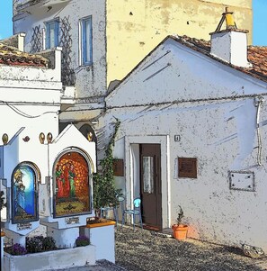 A narrow street with white buildings, a small shrine with religious icons, and a potted plant.
