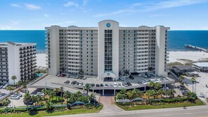 A multi-story hotel with a central entrance, parking area, and a view of the beach. - 🏖️ Admirals Quarters 1201 | 4BR Corner Condo | Gulf + Bay Views + Pool (Orange Beach)