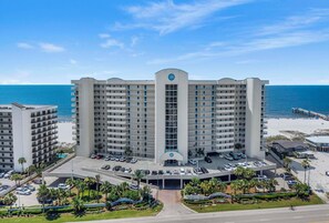 Ein mehrstöckiges Hotel mit zentralem Eingang, Parkplatz und Blick auf den Strand.