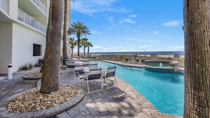 A pool area with lounge chairs, a hot tub, palm trees, and a building with balconies. - 🏖️ Admirals Quarters 1201 | 4BR Corner Condo | Gulf + Bay Views + Pool (Orange Beach)