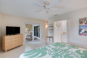 A bedroom with a TV, a ceiling fan, a bed with a green and white patterned bedspread, a small table, and a door leading to a balcony. - Villa Sanibel 3D | Pelican Perch Loft | Spacious Coastal Retreat (Sanibel)