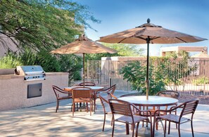 Une terrasse extérieure dotée d’un barbecue, de tables rondes et de chaises, le tout entouré d’arbres et d’une clôture.