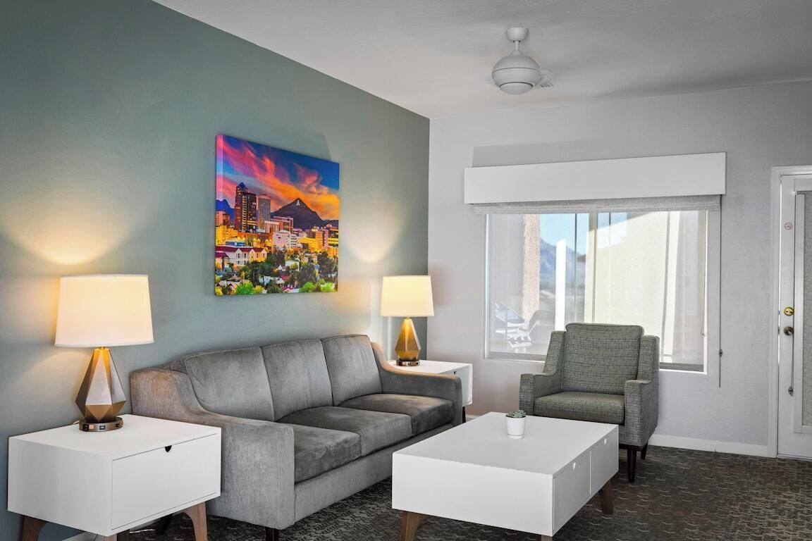 A hotel room with a gray sofa, a white coffee table, two side tables with lamps, a painting of a cityscape, and a window with a view of mountains.