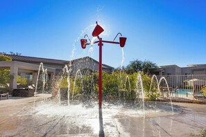 Una fuente con múltiples chorros, una piscina y un edificio al fondo.