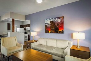 Habitación de hotel con sofá, sillón, mesa de centro y un cuadro de un atardecer en la pared.