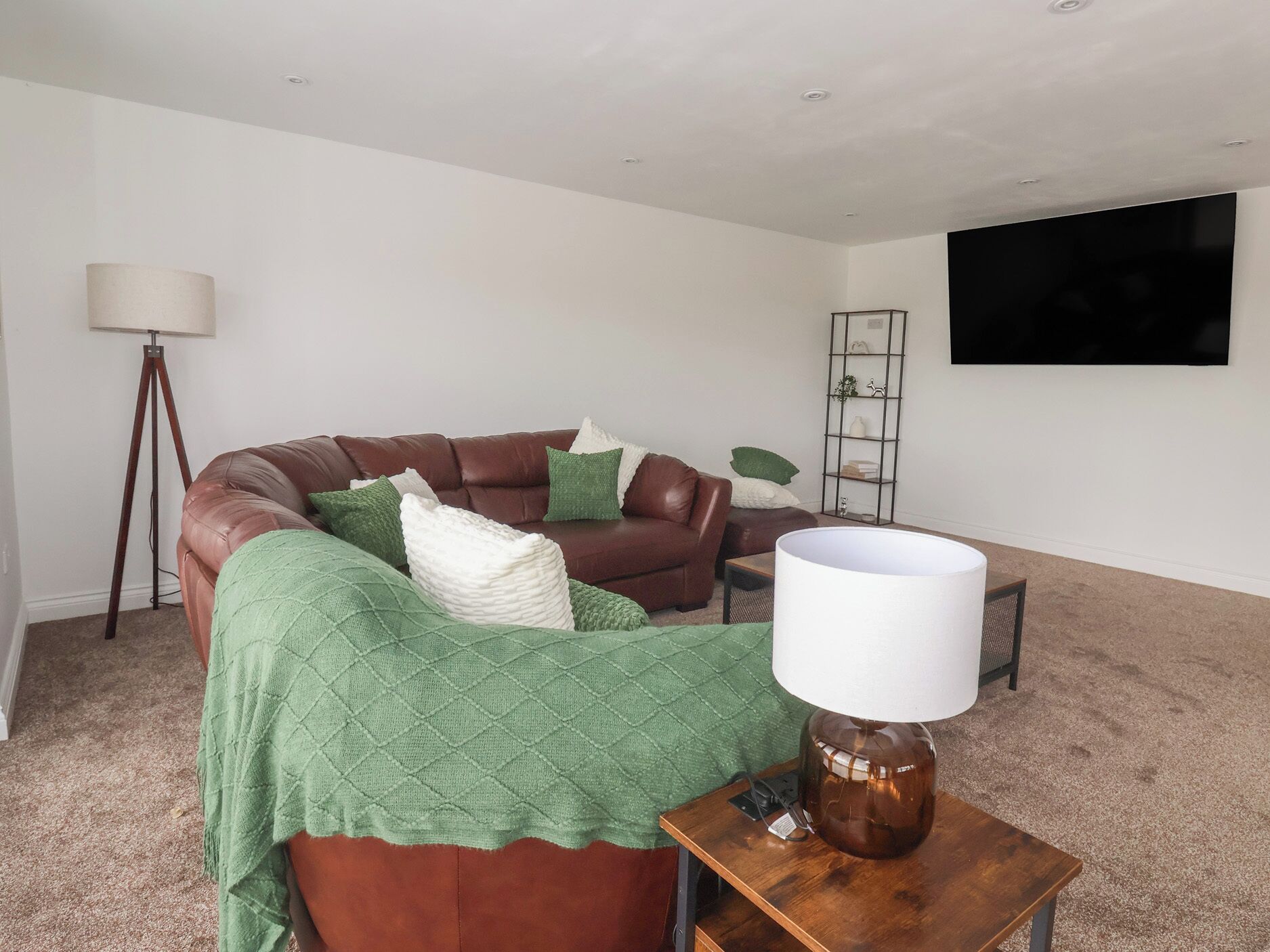 Un salón con un sofá de cuero marrón, una manta verde, una lámpara de pie, un televisor en la pared y una mesita con una lámpara.