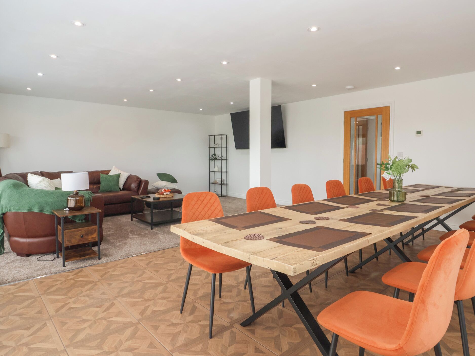 Uma sala de estar espaçosa com uma mesa comprida e cadeiras laranja, um sofá de couro marrom e uma televisão na parede.