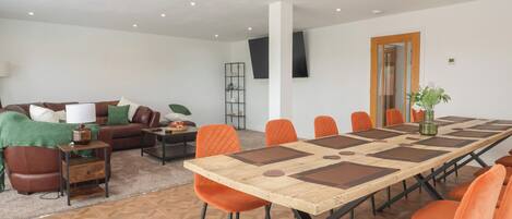 Uma sala de estar espaçosa com uma mesa comprida e cadeiras laranja, um sofá de couro marrom e uma televisão na parede.
