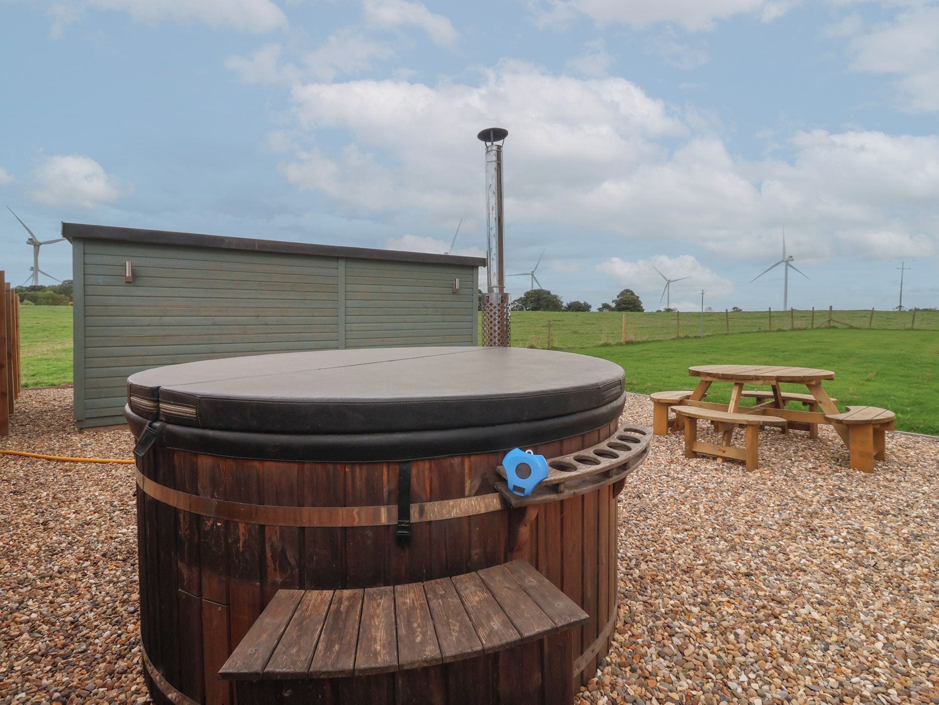 A hot tub with a wooden exterior, a wooden bench, and a picnic table in a gravel area with a green shed and wind turbines in the background.