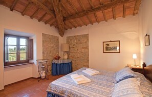 Um quarto com cama, janela, parede de pedra, luminária e quadro emoldurado.