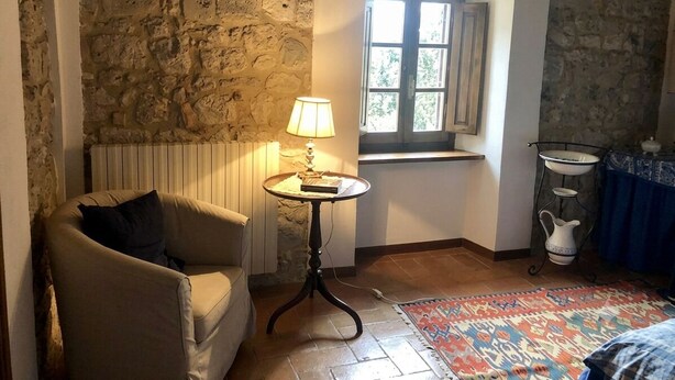Ett rum med en vägg av sten, ett fönster med gardiner, ett element, ett bord med en lampa, en soffa, en mönstrad matta och ett metallställ med en kanna.