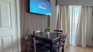 Zona pranzo con tavolo e sedie, televisione a parete che mostra Disney+, e una finestra con tende.